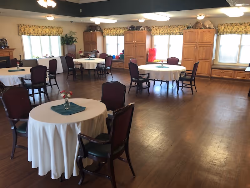 A spacious dining room with round tables covered in white tablecloths, each table surrounded by dark wooden chairs with cushioned seats. Small vases with flowers are placed on each table. The room has large windows with yellow floral valances, wooden cabinets along the walls, and a wooden floor.