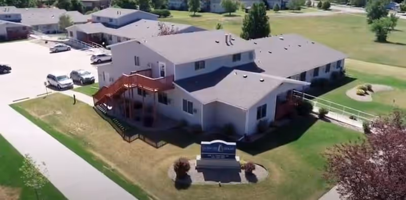Aerial view of a two-story senior living facility building with a parking lot, green lawns, trees, and a sidewalk. The building has exterior staircases and a sign in front indicating the facility name.