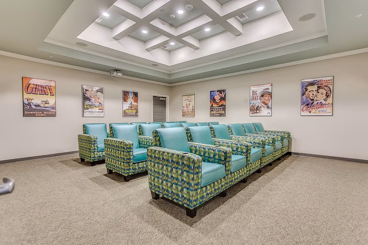 A small theater room with two rows of patterned armchairs featuring teal cushions, facing a blank wall with a projector mounted on the ceiling. The walls are decorated with vintage movie posters including titles like Giant, Casablanca, and Big Jake. The ceiling has a modern recessed lighting design.