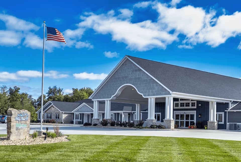 Exterior view of Villas of Holly Brook Assisted Living & Memory Care facility in East Peoria, IL, showing a large building with a covered entrance, an American flag on a flagpole, a stone sign, green lawn, and a partly cloudy blue sky.