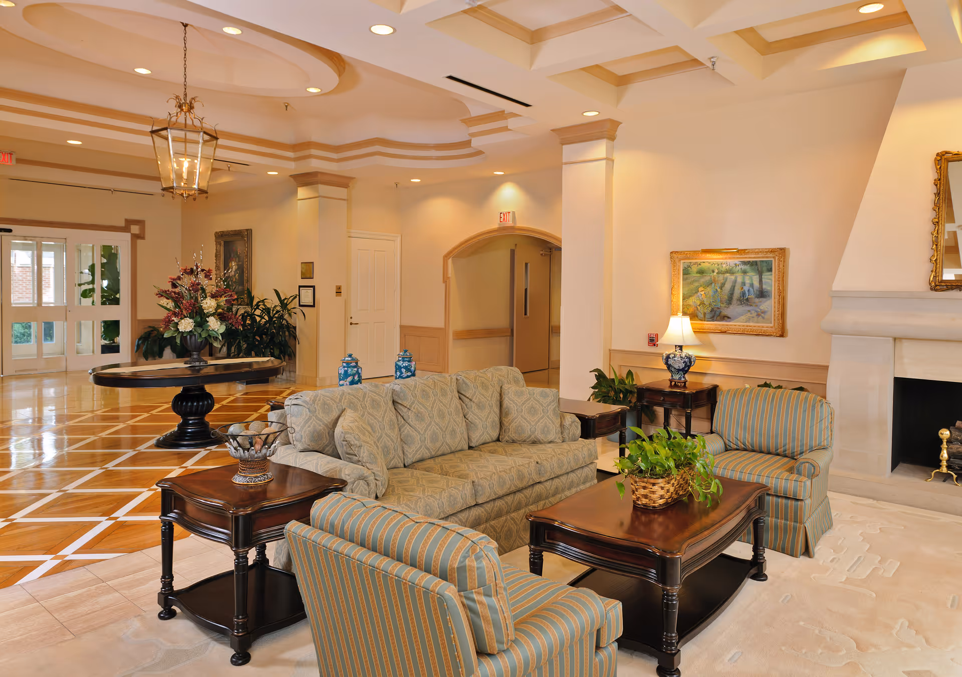 A spacious and elegant living room area in a nursing center featuring a patterned sofa and two striped armchairs around a wooden coffee table with a potted plant. The room has polished wooden floors with a geometric design, a large round table with a floral arrangement, a fireplace, framed artwork, and soft lighting from lamps and ceiling fixtures.