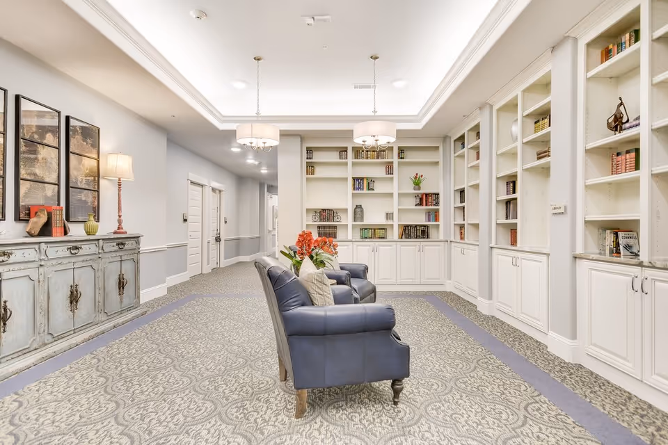 A bright and spacious senior living facility common area with two blue armchairs facing built-in white bookshelves filled with books and decorative items. The room features a patterned carpet, a vintage-style sideboard with a lamp and decor, and two hanging ceiling lights. The walls are painted light gray with white trim, and there are multiple doors leading to other rooms.