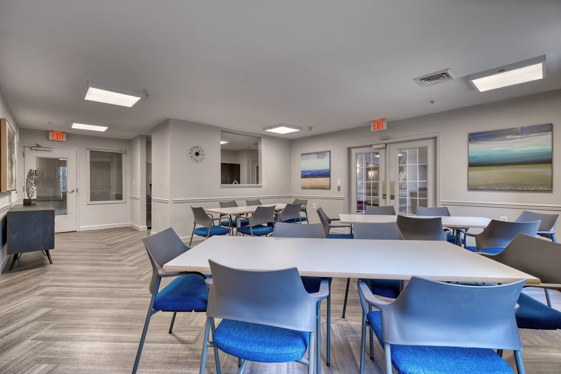 A bright and clean dining room with several white tables and gray chairs with blue cushions. The room has light gray walls, wood-patterned flooring, and modern ceiling lights. There are two exit signs above doors, a wall clock, and two abstract paintings on the walls.