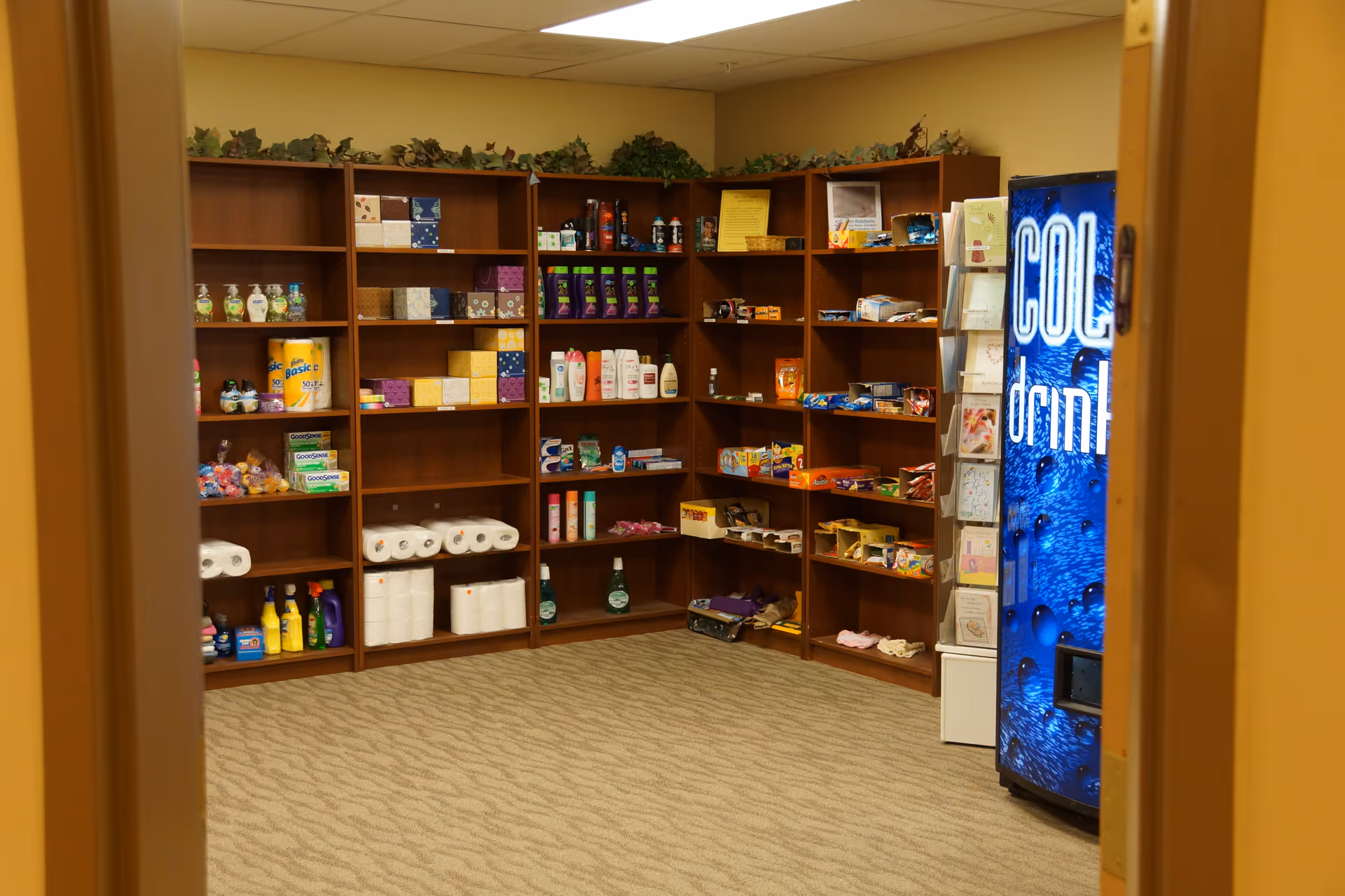 Small on-site shop or supply room with wooden shelves stocked with toiletries, snacks and household items and a blue beverage vending machine on the right.