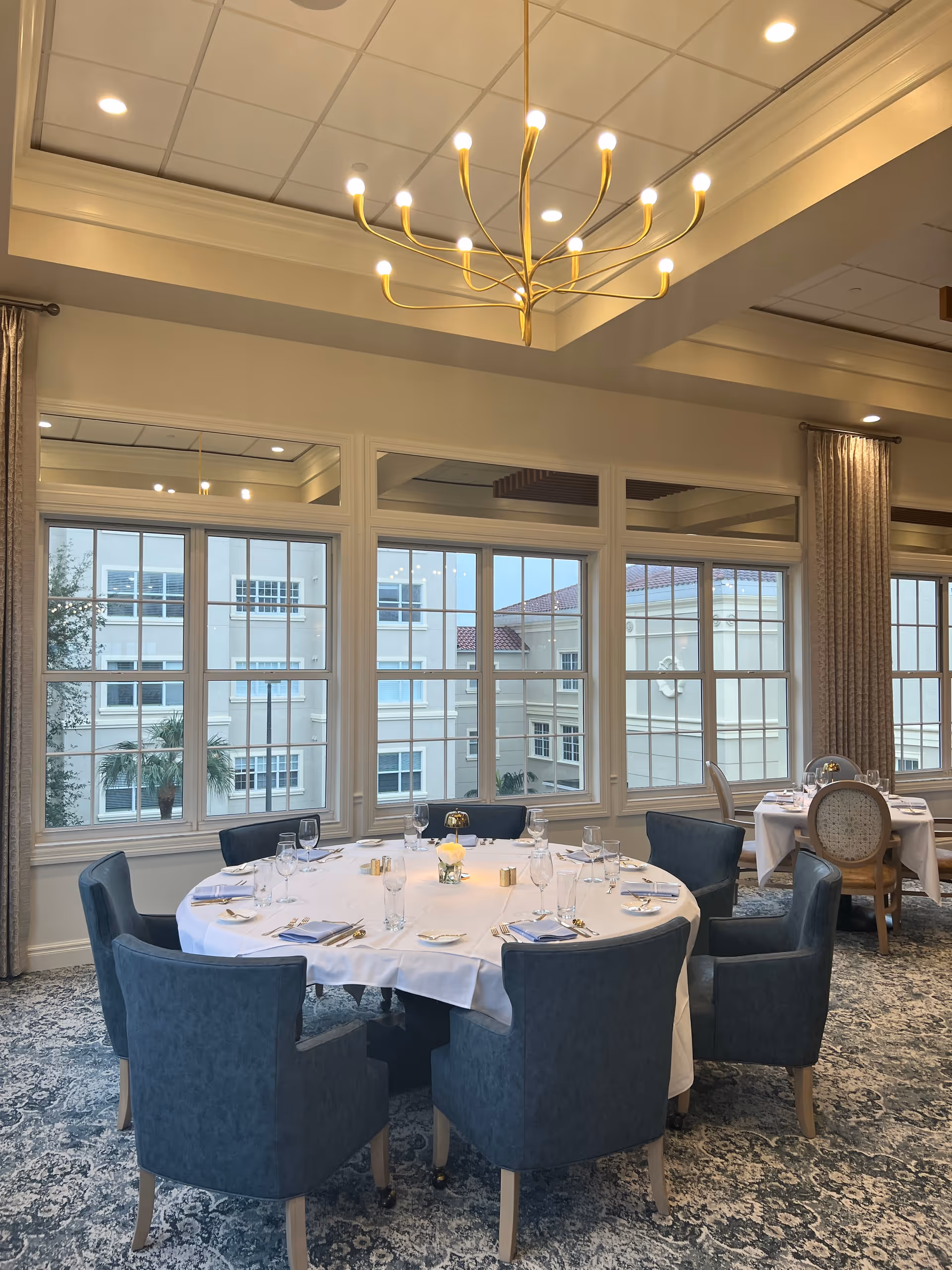 A dining room with a round table set for a meal, surrounded by six blue upholstered chairs. The table is covered with a white tablecloth and set with glassware, napkins, and cutlery. Large windows with a view of an adjacent building allow natural light to fill the room. A modern chandelier hangs from the ceiling, and curtains frame the windows.