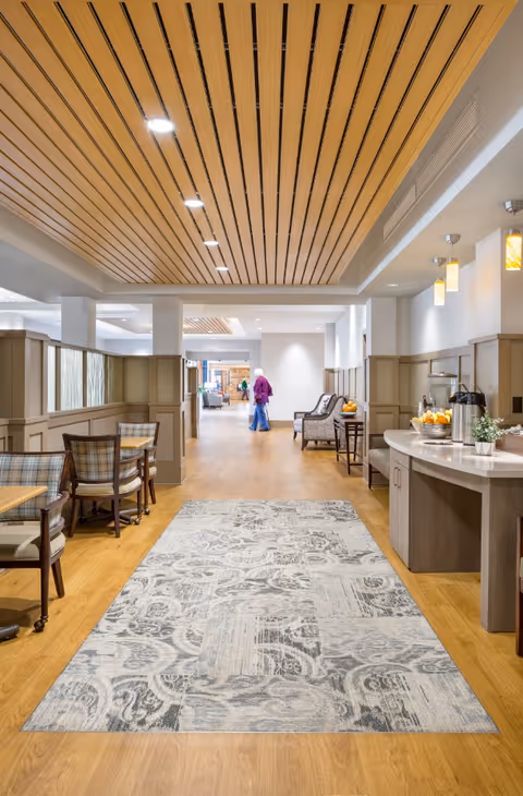 A spacious, well-lit interior hallway in a senior living facility with wooden flooring and a patterned area rug. On the left side, there are tables and chairs with plaid upholstery. On the right side, there is a counter with coffee dispensers, cups, and a small plant. The ceiling features wooden slats with recessed lighting. In the background, a person wearing a purple top and blue pants is walking away from the camera.