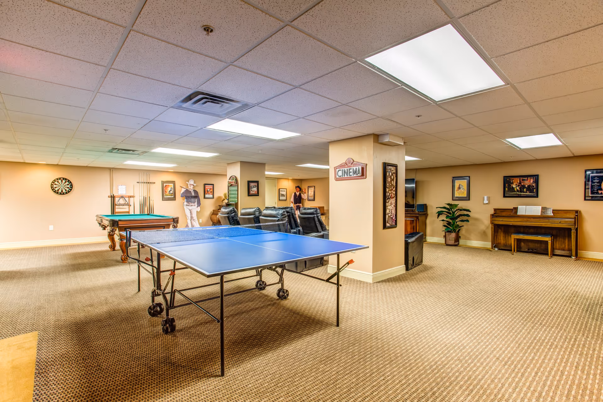 A spacious recreational room with a ping pong table in the foreground, a pool table and dartboard on the left wall, several black leather chairs arranged in front of a TV, a piano on the right side, and various framed pictures and a plant decorating the beige walls.