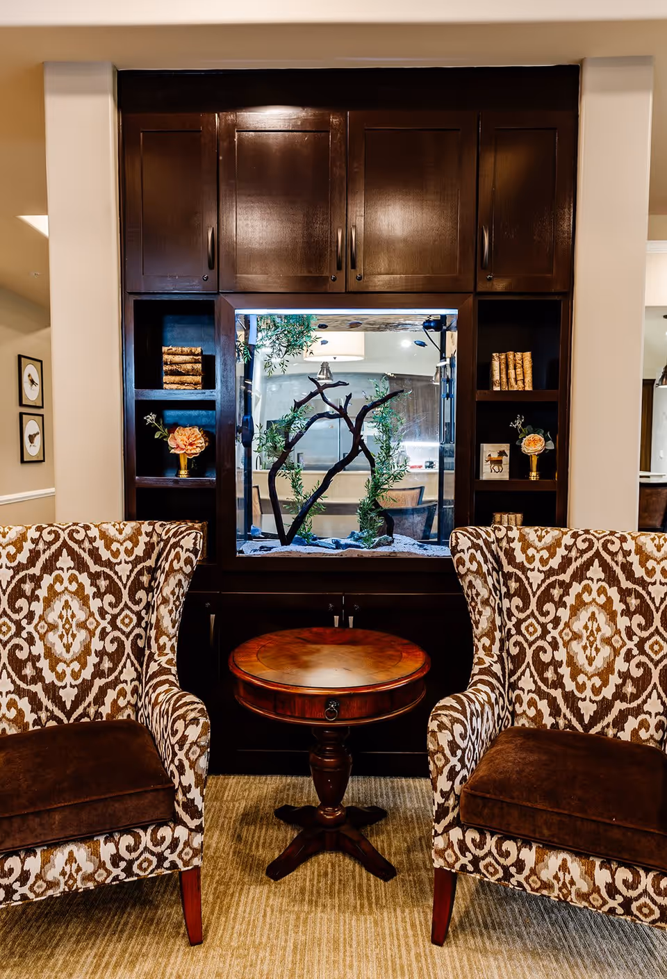 Two patterned armchairs with brown cushions are positioned on either side of a small round wooden table. Behind the table and chairs is a built-in dark wood cabinet with an aquarium in the center, decorated with branches and greenery. Shelves on either side of the aquarium hold decorative items including flowers and books. The setting appears to be a cozy indoor seating area.