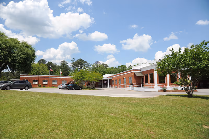 Single-story red brick senior living facility building with a parking area, flagpoles, a wheelchair ramp, and a grassy lawn under a blue sky.
