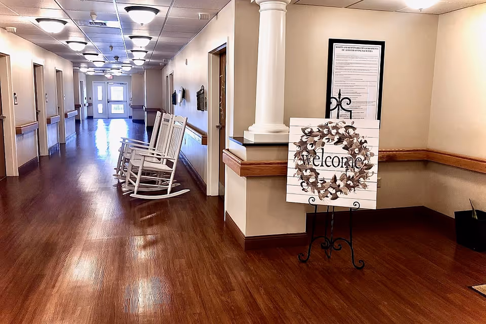 A long, well-lit hallway with wooden flooring and beige walls. There are white rocking chairs lined up along one side of the hallway. At the near end, a decorative sign on an easel reads 'welcome' with a wreath design. A framed notice is mounted on the wall behind the sign. The hallway has multiple doors and handrails along the walls.