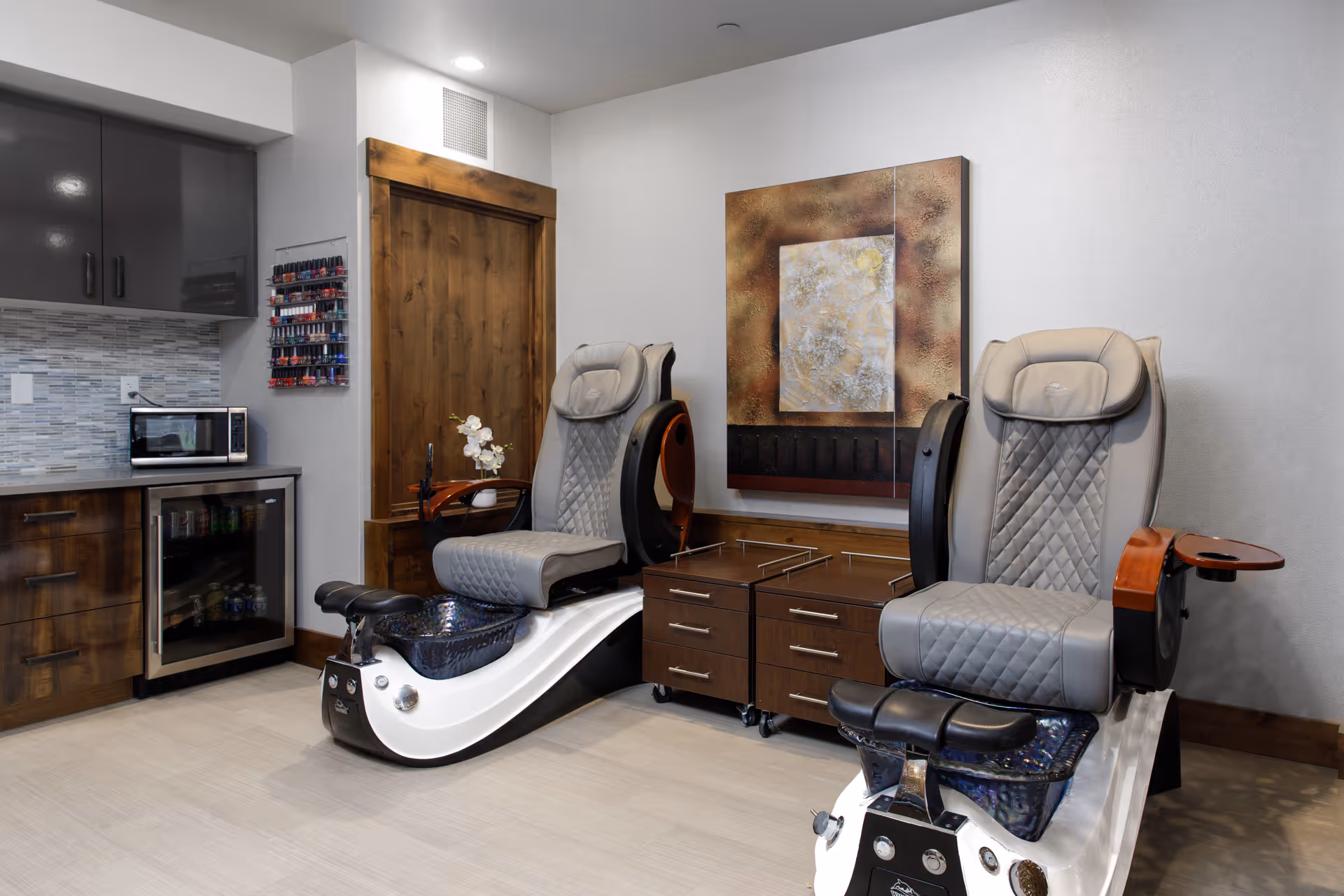 A modern nail salon area with two gray cushioned pedicure chairs featuring foot baths. The room has wooden cabinetry, a small refrigerator, a microwave, and a wall-mounted rack with various nail polish bottles. There is a decorative abstract painting on the wall behind the chairs.