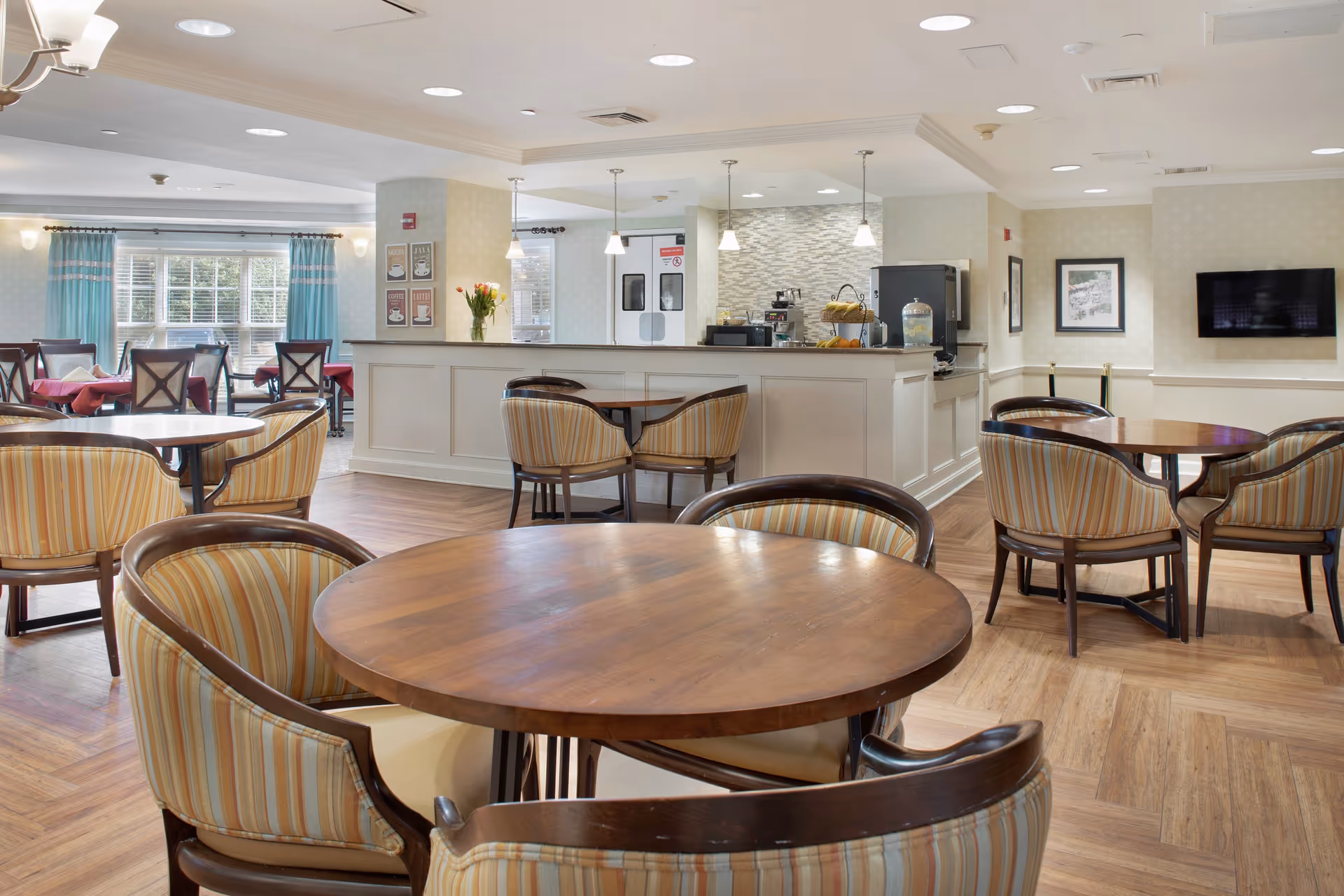 Bright dining/lounge area with round wooden tables and striped upholstered chairs facing a service counter and wall-mounted TV.