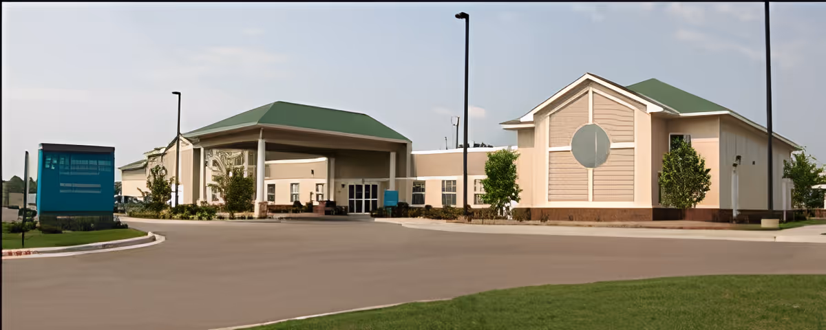 Exterior view of a senior living facility showing the building front with a covered porte-cochere, driveway, and landscaped grounds.