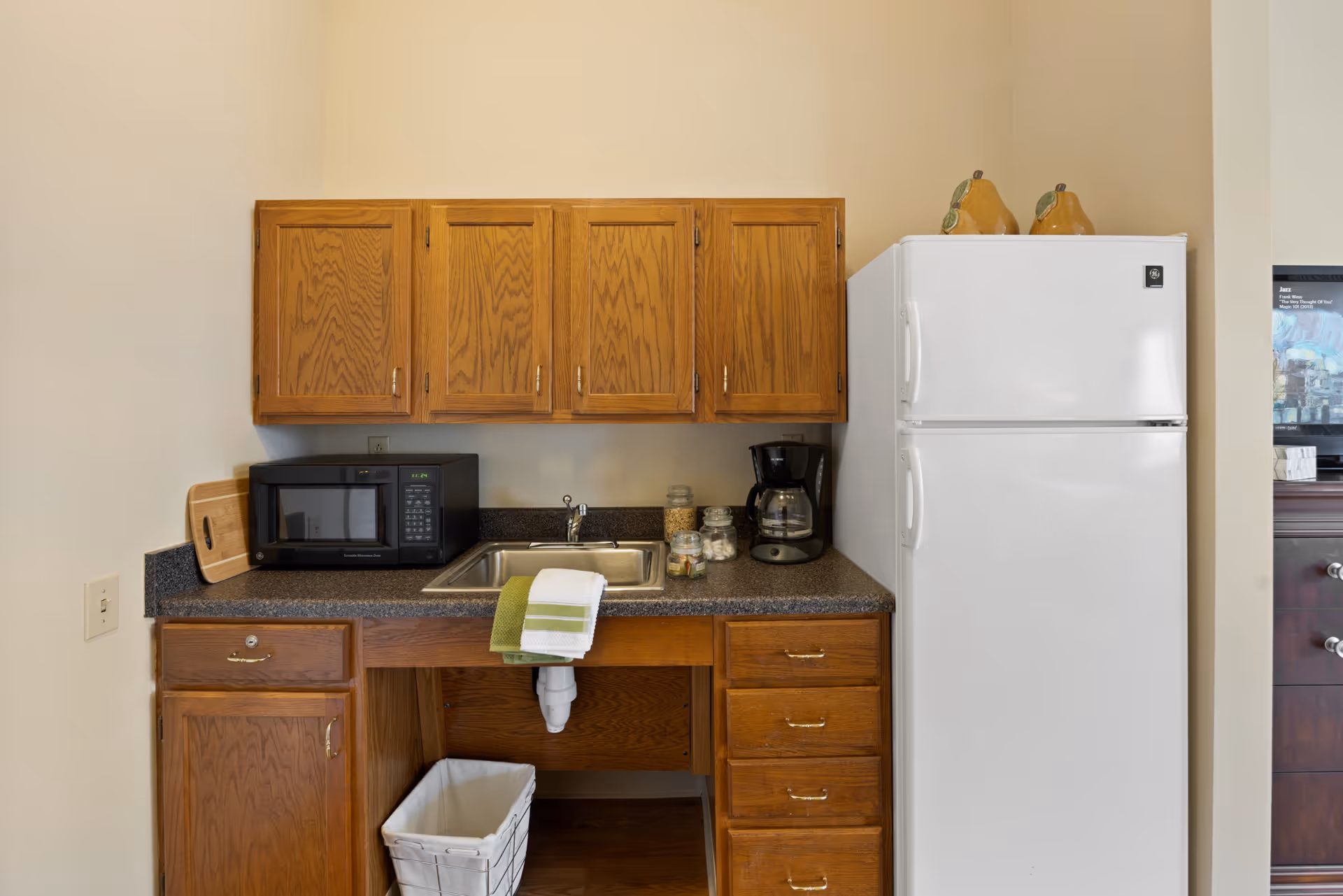 A small kitchen area with wooden cabinets, a black microwave, a stainless steel sink with a green and white towel hanging over the edge, a coffee maker, and a white refrigerator. There are two decorative pear-shaped items on top of the refrigerator and a white trash bin under the sink.