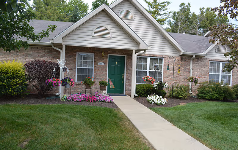 Exterior view of a single-story brick building with a green front door, white-framed windows, and a concrete walkway leading to the entrance. The front yard is landscaped with green grass, bushes, and colorful flower arrangements in pots and hanging baskets.
