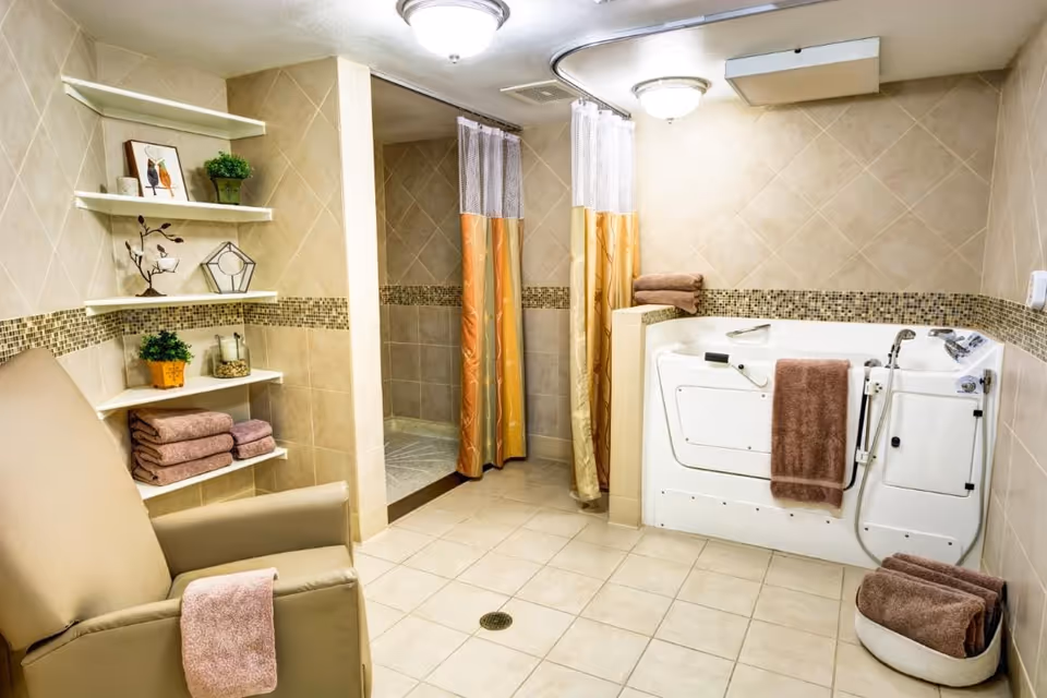 A spacious bathroom with beige tiled walls and floor, featuring a walk-in shower with two orange and yellow shower curtains, a white walk-in bathtub with brown towels hanging on it, a beige armchair with a brown towel draped over one arm, and white corner shelves holding decorative items and folded towels.