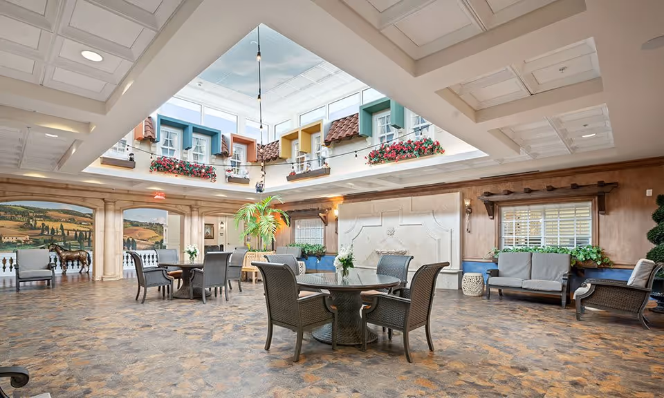 Spacious indoor common area with multiple seating arrangements including round tables with chairs and cushioned benches. The ceiling features a skylight with decorative window boxes and flower arrangements. The walls have a mural of a countryside landscape and architectural details resembling columns and windows.