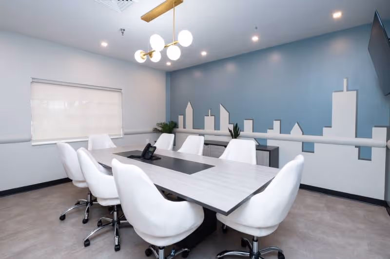 A modern conference room with a rectangular table surrounded by eight white swivel chairs. The room has a blue accent wall featuring a white city skyline design, a window with a closed white blind, a ceiling light fixture with round bulbs, and a wall-mounted TV.