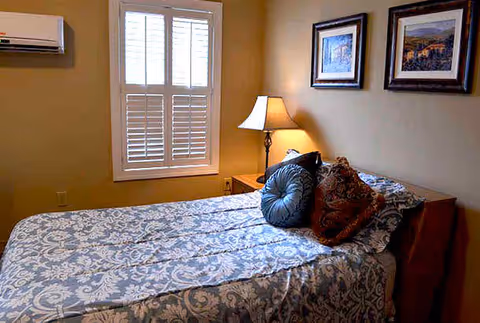 Cozy bedroom with a patterned bedspread, decorative pillows, a bedside lamp and framed art under a window with shutters.