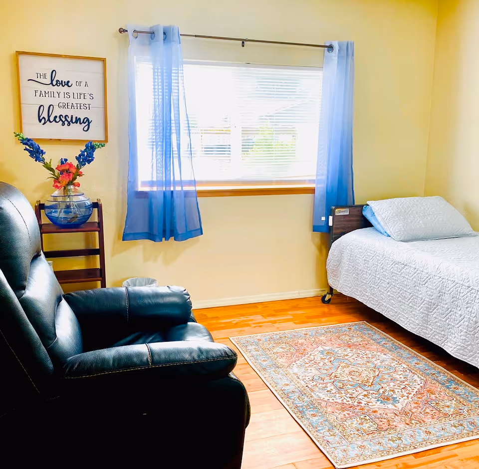 A cozy bedroom with a single bed covered in a white quilt, a black leather recliner chair, a colorful patterned rug on a wooden floor, and a window with blue sheer curtains letting in natural light. A small wooden stand with a glass vase holding blue and pink flowers is next to the chair, and a framed wall sign reads 'The love of a family is life's greatest blessing.'