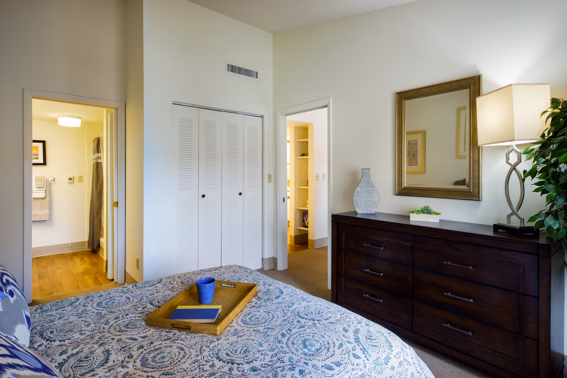 A bedroom with a bed covered in a blue and white patterned bedspread. On the bed is a wooden tray holding a blue cup and a book. To the right is a dark wooden dresser with a decorative vase, a small plant, and a lamp with a beige shade. Above the dresser is a large framed mirror. There is a doorway leading to a bathroom with towels hanging on the wall and a shower curtain visible. Another doorway leads to a hallway with shelves. The room has light-colored walls and carpeted floors.