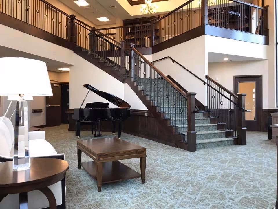 Interior view of a senior living facility featuring a grand staircase with dark wood railings and green carpeted steps. A black grand piano is positioned near the staircase. The room includes a white sofa, a wooden coffee table, and a side table with a modern lamp. The space is well-lit with ceiling lights and has a high ceiling with a chandelier.
