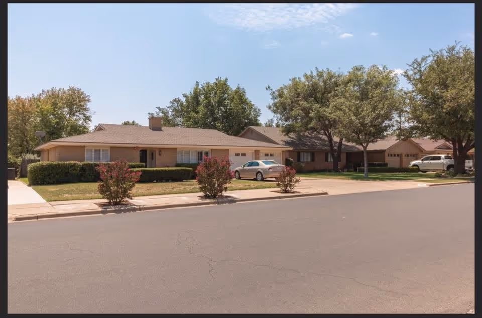 Single-story ranch-style building with a front lawn, trees, and parked cars along a quiet residential street.
