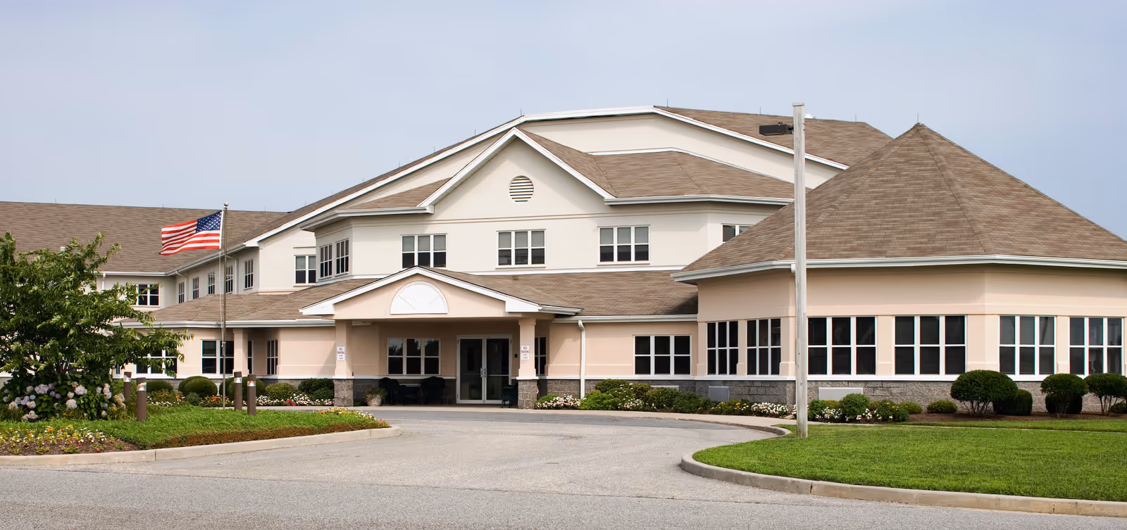 Front exterior of a two-story senior living facility with a covered entrance, circular driveway, landscaped lawn, and an American flag.