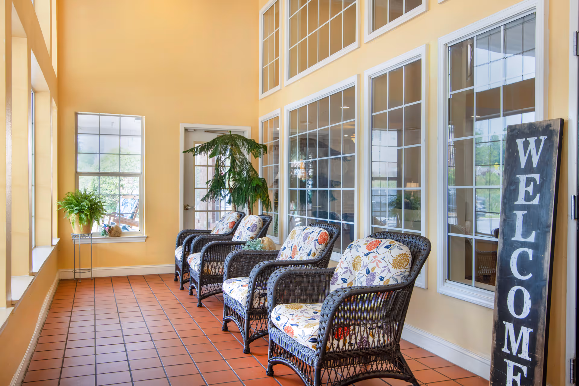 A bright indoor seating area with four wicker chairs featuring floral cushions arranged in a row along a wall with large windows. There are two potted plants, one on a stand and one on the floor, and a tall black sign with the word 'WELCOME' leaning against the wall. The floor is tiled with reddish-brown tiles and the walls are painted a warm yellow.