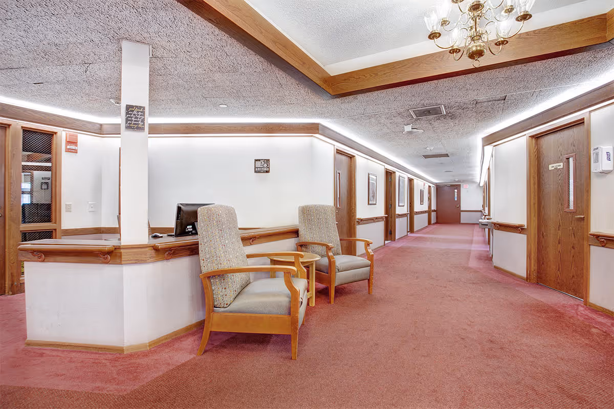 Interior view of a senior living facility hallway with a reception desk on the left side, two cushioned wooden armchairs in front of the desk, and several closed wooden doors along the right side. The hallway has a red carpet, white walls with wooden trim, framed pictures on the walls, and a chandelier hanging from the ceiling.