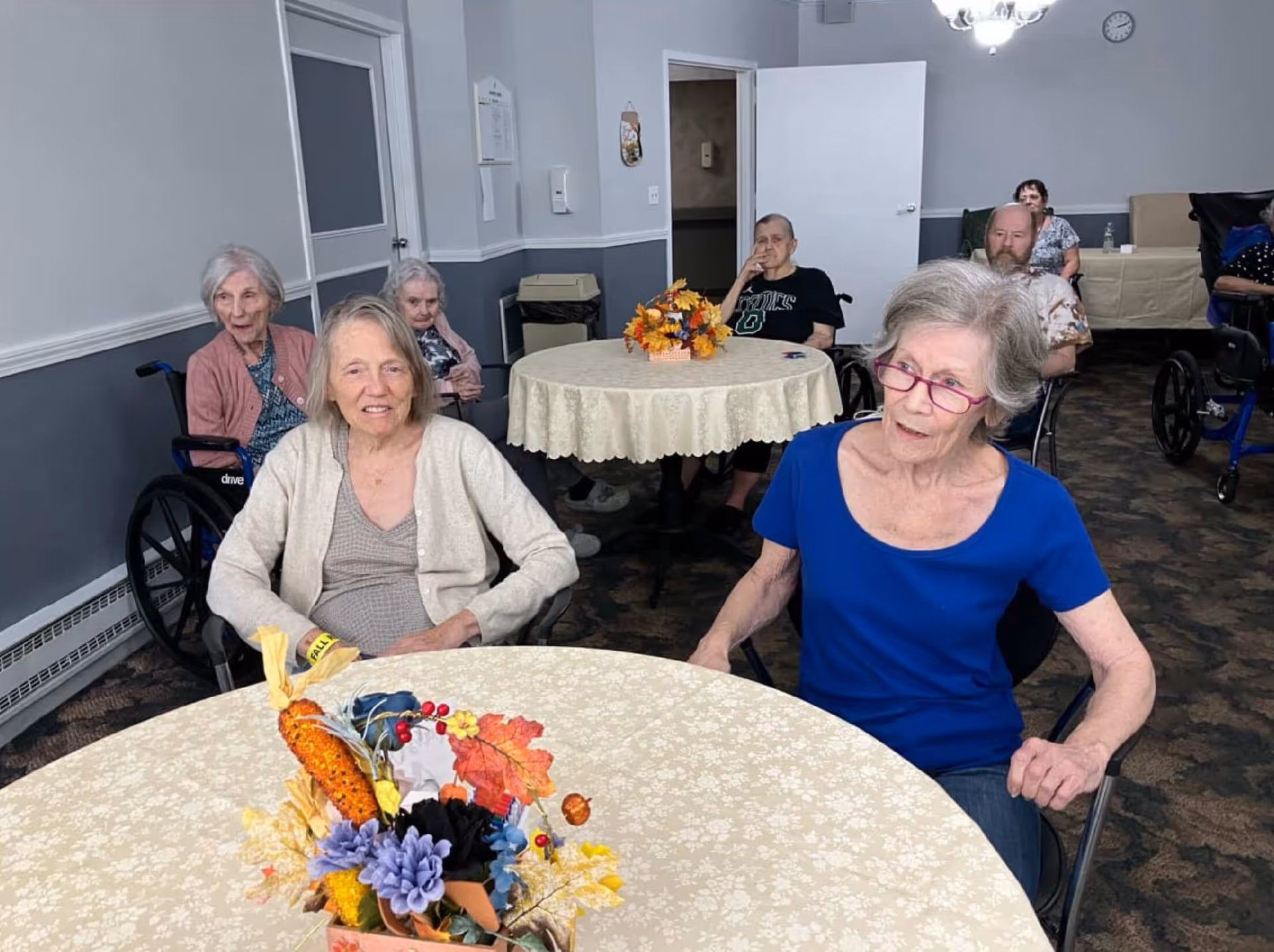A group of elderly individuals sitting around tables in a common room. Two women are seated at the front table with a floral centerpiece, while others are seated at tables and in wheelchairs in the background. The room has light gray walls, a carpeted floor, and a clock on the wall.