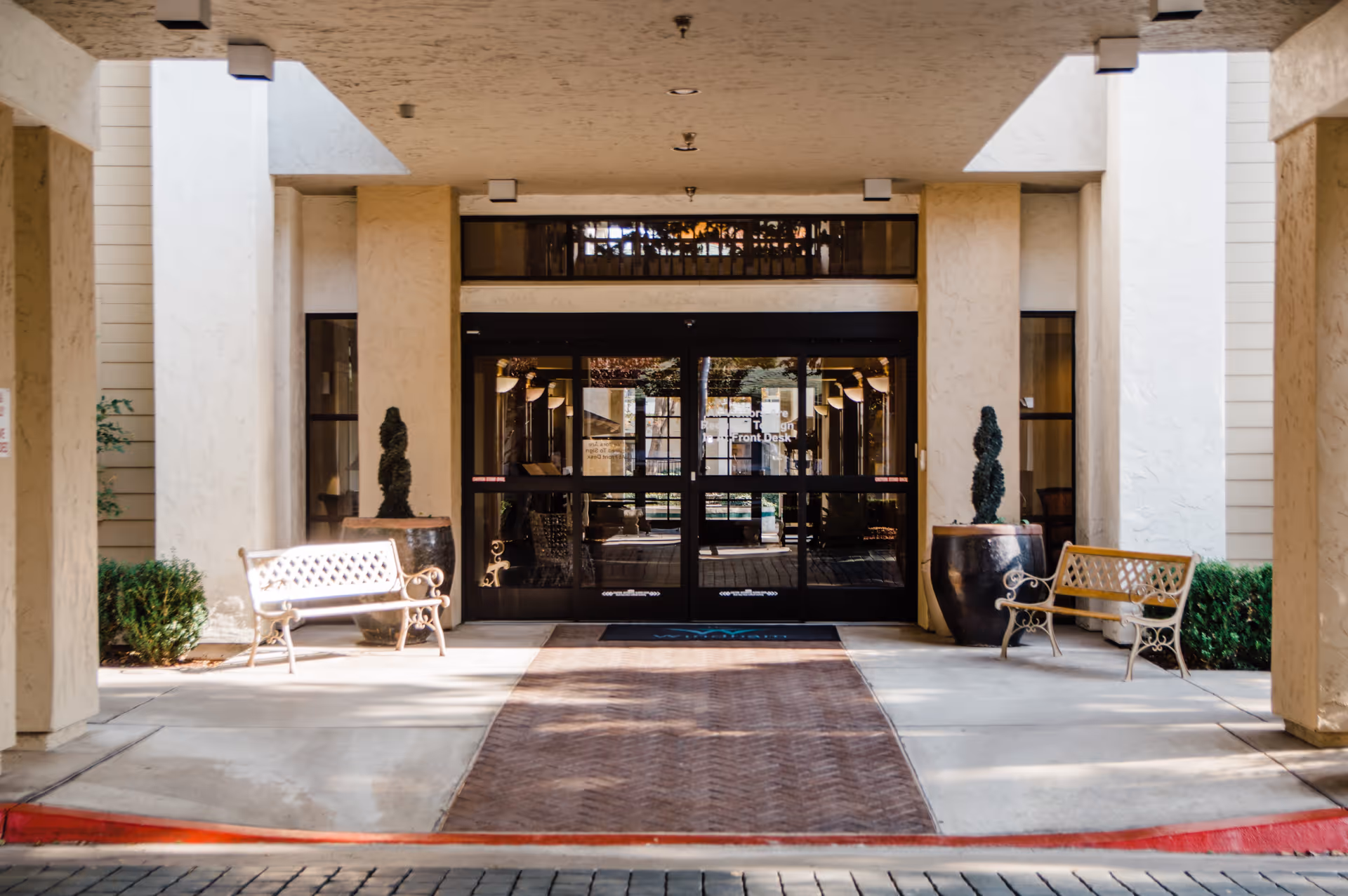 Entrance to a building with double glass doors, flanked by two large potted plants and two metal benches on either side. The area is covered by a ceiling with recessed lighting and has a brick-patterned walkway leading to the doors.