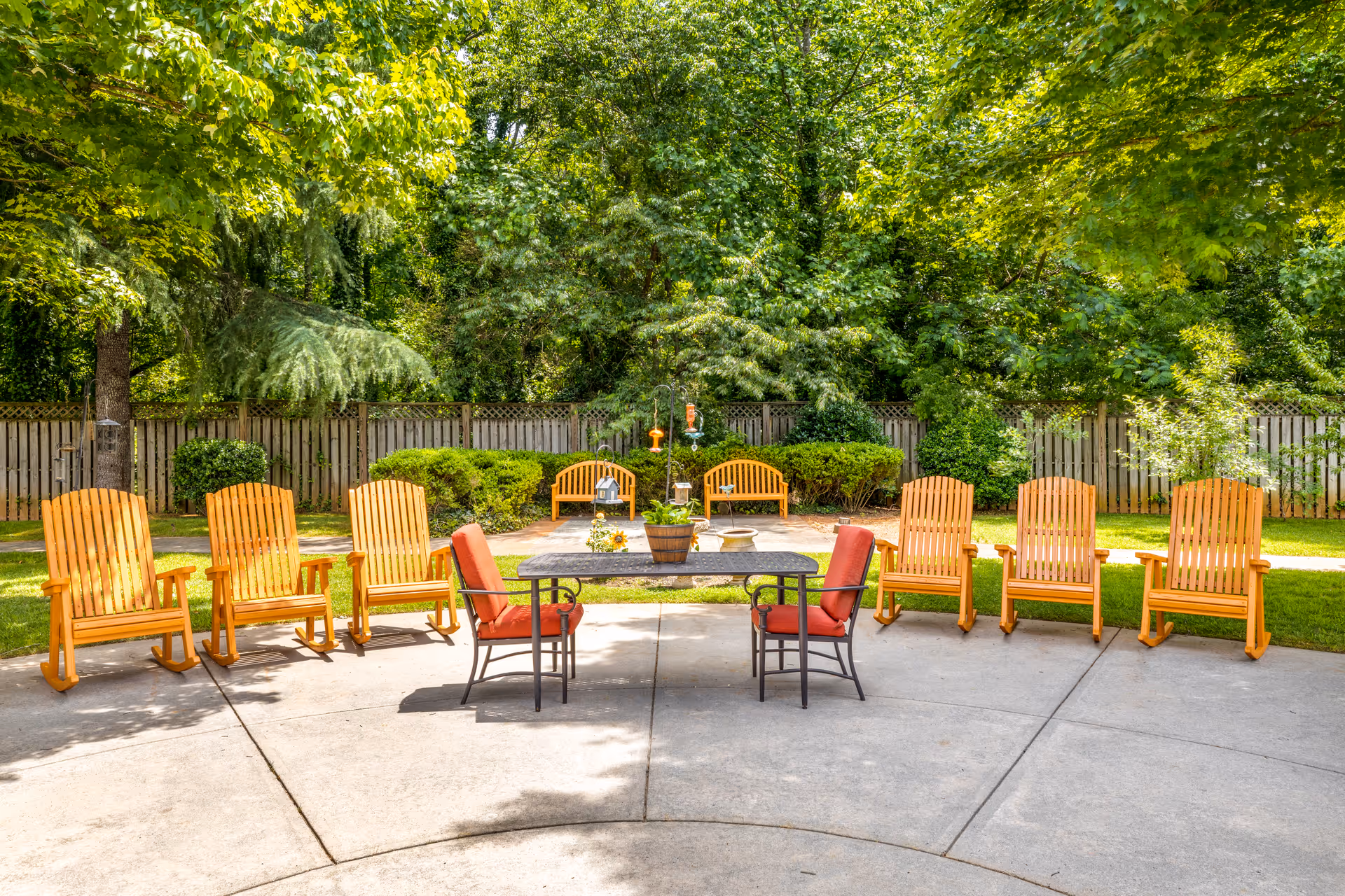 Outdoor patio area with a semi-circle arrangement of wooden Adirondack chairs and two red cushioned chairs around a rectangular table, surrounded by green trees and bushes with a wooden fence in the background.