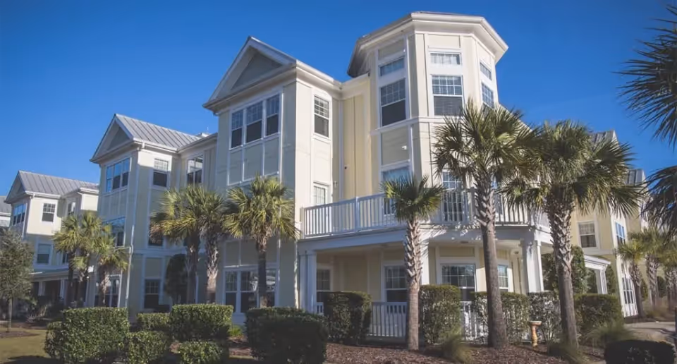 Exterior view of a multi-story senior living facility building named Brightwater, featuring large windows, balconies, palm trees, and well-maintained landscaping under a clear blue sky.