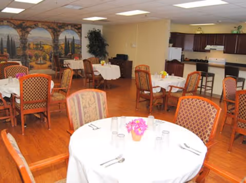 Bright communal dining room with round tables set with white tablecloths and chairs, a kitchenette along the back wall and a mural on the left wall.