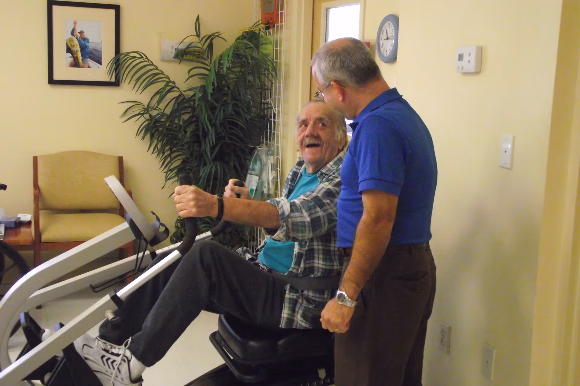 An elderly man is sitting on a seated exercise machine, holding the handles and smiling while looking at another man standing next to him who is wearing a blue shirt and glasses. The room has light yellow walls, a clock on the wall, a thermostat, a framed picture, a potted plant, and a beige chair in the background.