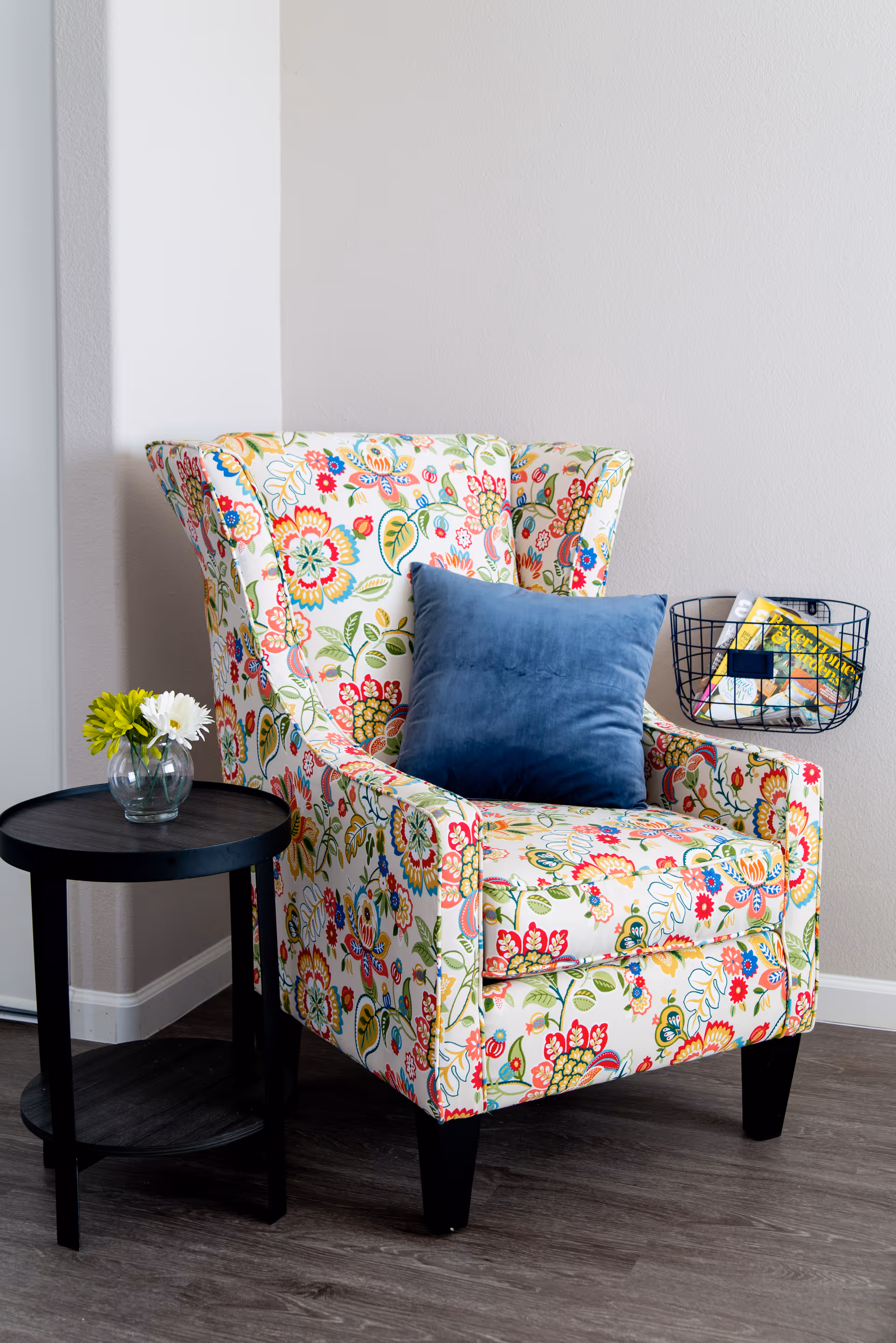Colorful floral upholstered armchair with a blue throw pillow next to a black side table with a small vase of flowers and a wire basket of magazines.