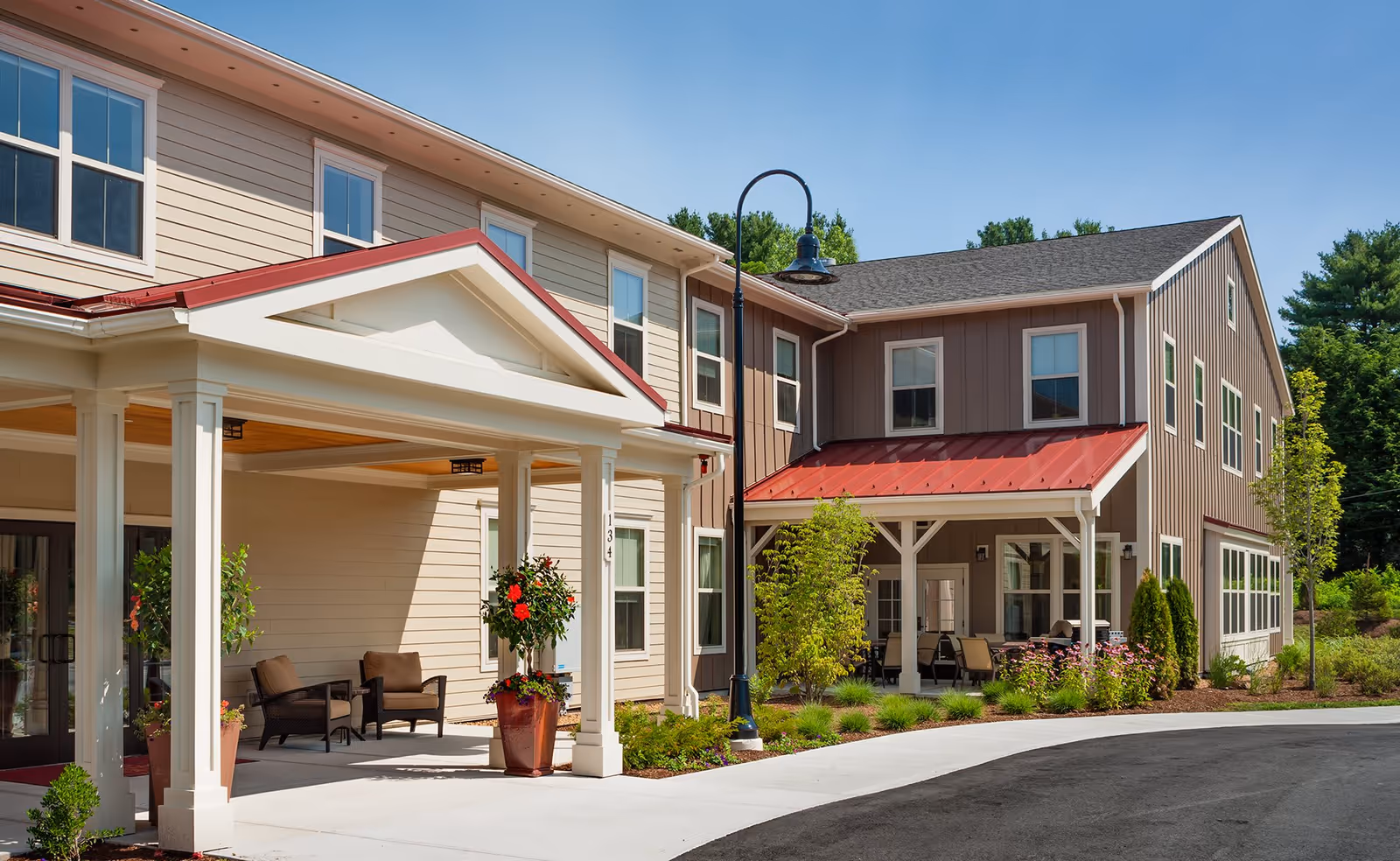 Exterior view of a senior living facility building with beige and brown siding, red metal roofs over entrances, white columns, potted plants, outdoor seating areas, and a curved driveway under a clear blue sky.