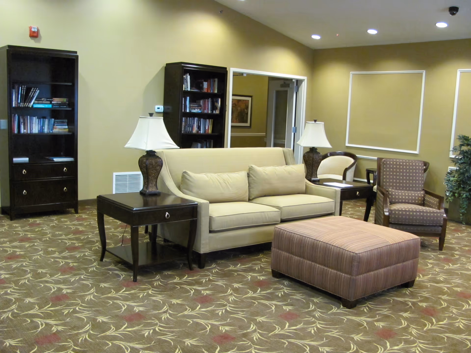 A cozy living room area with a beige sofa, two dark wood side tables with lamps, a striped ottoman, two armchairs, two dark wood bookshelves filled with books, and a patterned carpet. The walls are painted a warm beige color with white trim and there is a doorway leading to another room.