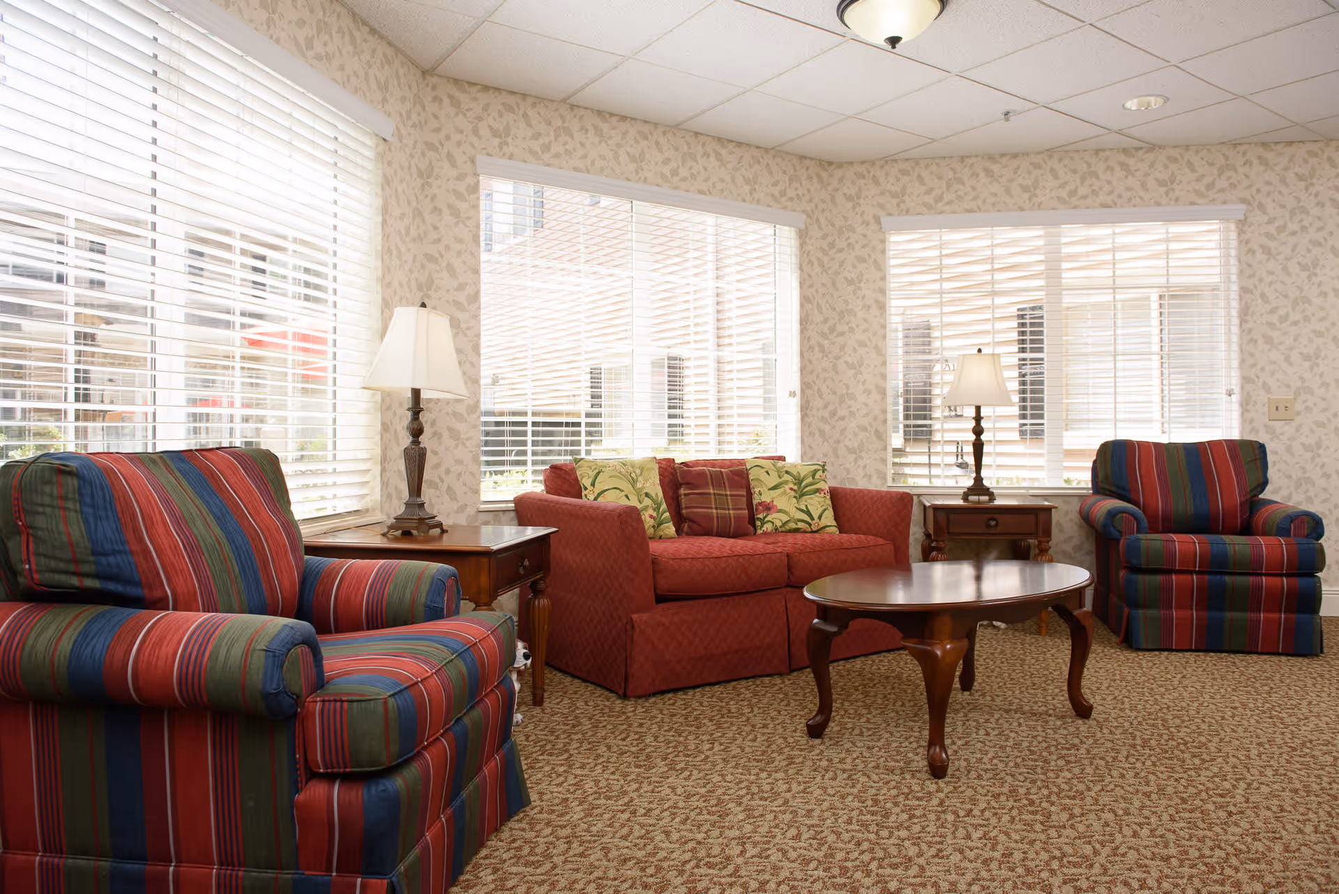 A cozy living room with two striped armchairs and a red sofa adorned with patterned cushions. The room features large windows with white blinds, two wooden side tables each holding a lamp, and a wooden coffee table in the center. The walls have a light floral wallpaper and the floor is carpeted with a patterned design.