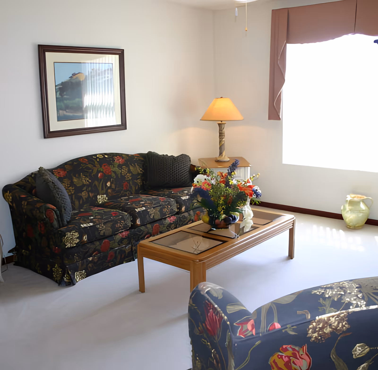 A bright living room with floral patterned sofas, a wooden coffee table with a glass top holding a colorful flower arrangement, a side table with a lamp, a large window with a valance, a framed picture on the wall, and a ceramic vase on the floor.