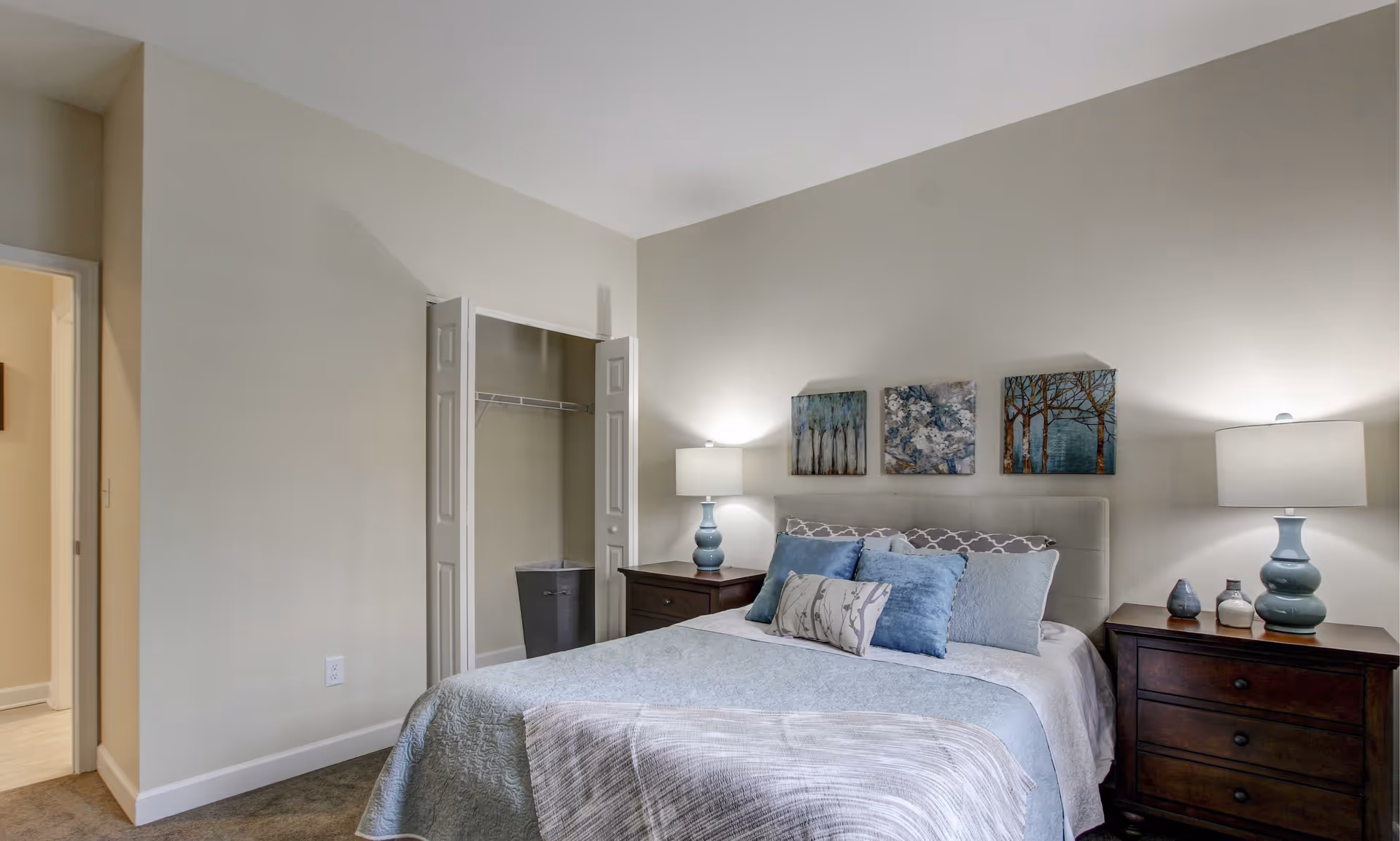 A bedroom with a neatly made bed featuring blue and gray pillows and a light blue quilt. There are two dark wooden nightstands on either side of the bed, each with a blue lamp. Above the bed, three framed artworks depicting trees and floral designs are hung on the light-colored wall. An open closet with white bi-fold doors and a laundry basket inside is visible to the left of the bed. The room has beige walls and carpeted flooring.