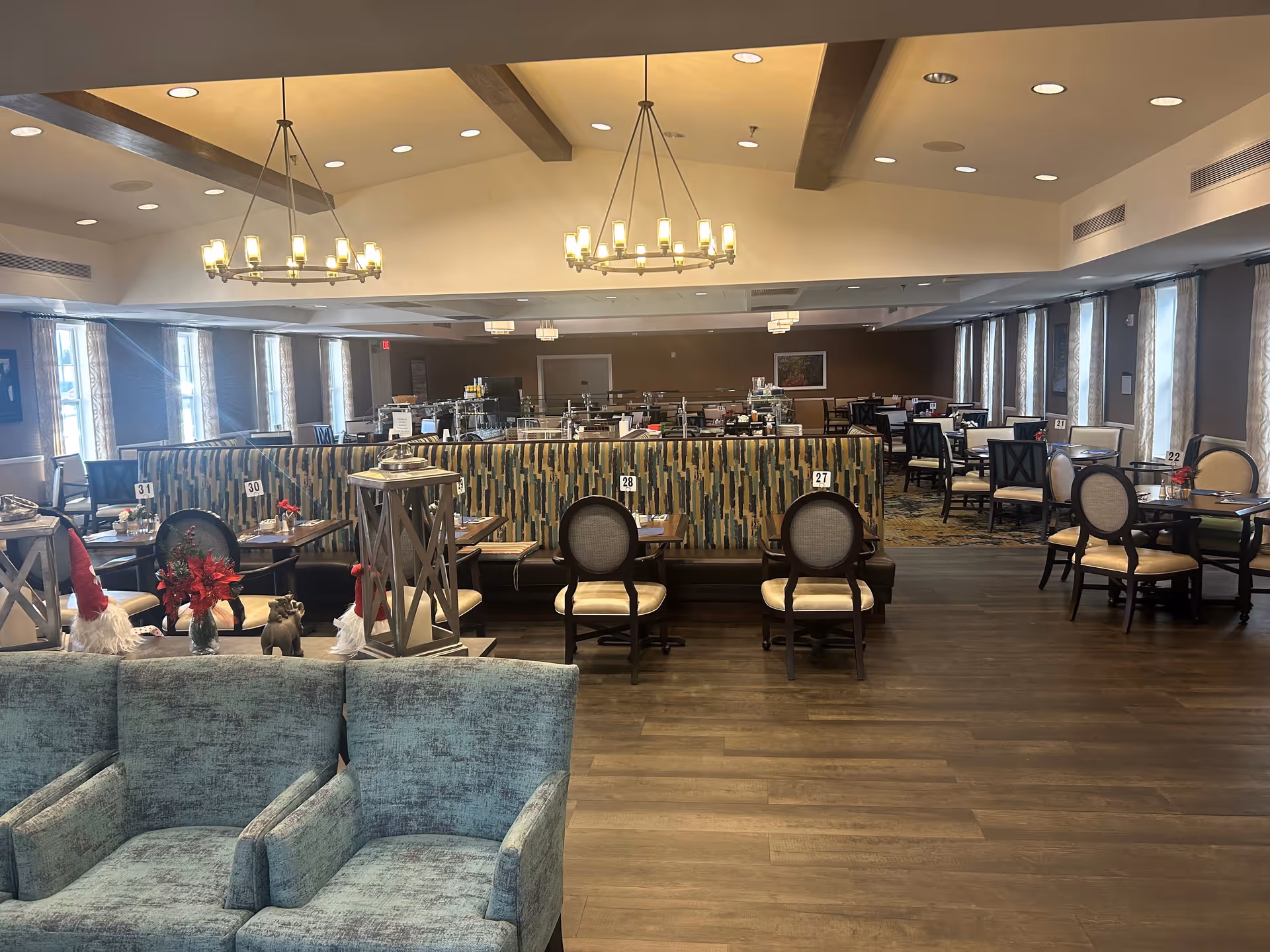 A spacious dining room in a senior living facility with multiple tables and chairs arranged neatly. The room features large windows with curtains, modern chandeliers hanging from a ceiling with wooden beams, and a patterned booth seating area in the center. The floor is wooden, and there are decorative items including small holiday-themed figurines on a ledge in the foreground.