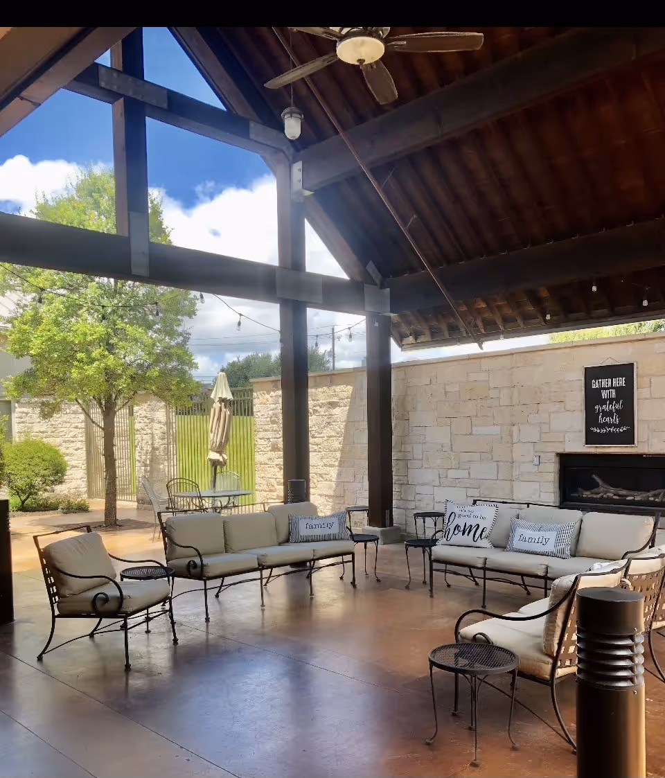 Covered outdoor patio with cushioned metal lounge seating, a fireplace, and large wooden beams under a pitched roof.