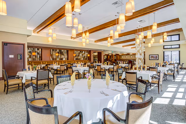 A spacious, well-lit dining room with multiple round and rectangular tables covered with white tablecloths. Each table is set with folded yellow napkins, glassware, and silverware. The room features wooden ceiling beams, modern hanging pendant lights, large windows letting in natural light, and comfortable chairs arranged around the tables.
