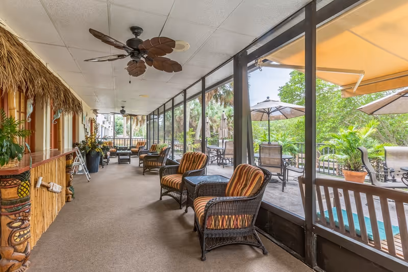 A screened-in porch area with wicker chairs featuring striped cushions, small tables, ceiling fans with leaf-shaped blades, and a tiki-style bar on the left. Outside the screened area, there are patio tables with umbrellas and lush green trees in the background.