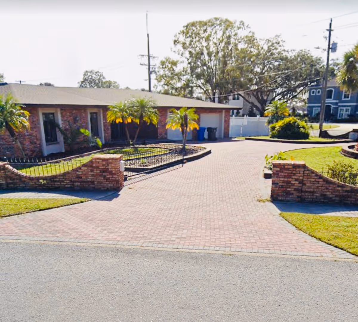 Front exterior view of a single-story brick building with a paved driveway, small palm trees, and landscaped garden areas enclosed by low brick walls.