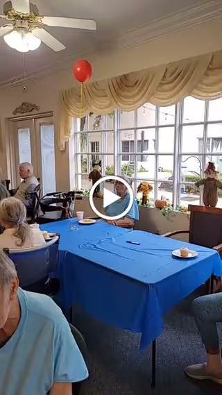 Several seniors seated around a table with a blue tablecloth and snacks in a bright common room with large windows, plants, and a red balloon.