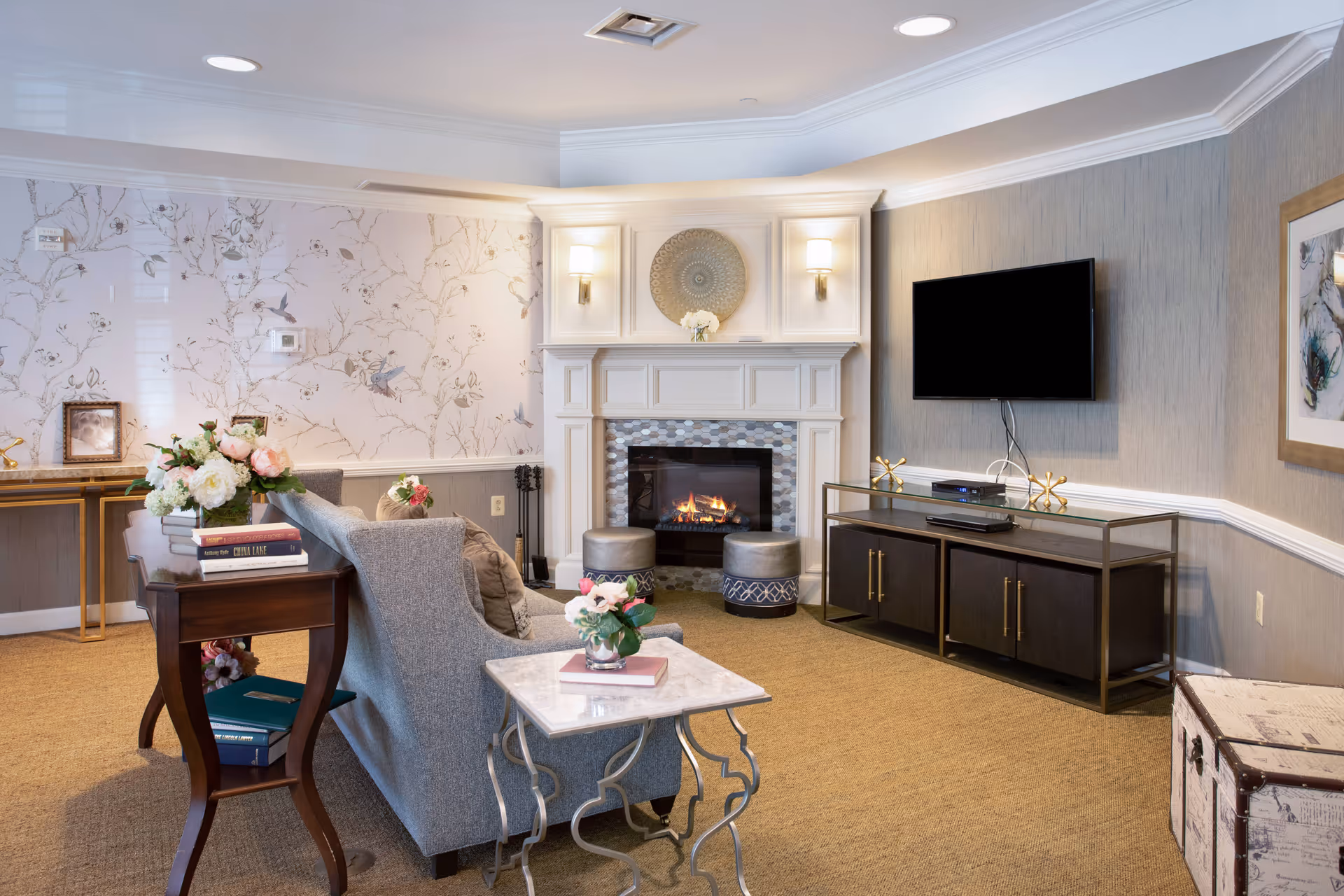 A cozy living room with a gray sofa facing a white fireplace with a fire burning. The room features a mounted flat-screen TV on the wall, a dark wood TV console, a small marble-topped side table with a flower vase, and a wooden side table with books and flowers. The walls have a combination of floral wallpaper and textured gray paint, and the room is warmly lit with wall sconces and natural light.