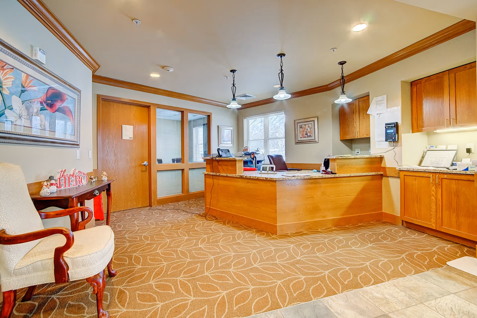 Reception area with a wooden desk, office chair, and cabinets. The room has beige walls, a patterned carpet, and three hanging pendant lights. There is a wooden door and windows with blinds behind the desk. A cushioned armchair and a small table with decorative items and a 'thank you HERO' sign are visible on the left side. A framed floral painting hangs on the wall.